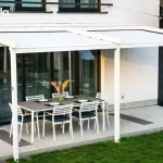 Szeroka, biała pergola tarasowa Tarasola Simple o konstrukcji przyściennej, zamontowana na elewacji nowoczesnego domu jednorodzinnego. Zadaszenie składa się z dwóch sekcji białego, napiętego materiału technicznego, zamocowanego na aluminiowych profilach poprzecznych i prowadnicach. Konstrukcja opiera się na trzech pionowych słupach o kwadratowym przekroju, które są zakotwiczone w gruncie, na trawniku przy krawędzi tarasu. Pod zadaszeniem znajduje się strefa wypoczynkowa z dużym, prostokątnym stołem o szarym blacie oraz sześcioma białymi krzesłami z ciemnymi poduszkami. Taras został wyłożony jasnymi, nakrapianymi płytami z ozdobnymi ciemniejszymi pasami. Elewacja budynku łączy biały tynk z panelami imitującymi naturalne drewno w ciepłym brązowym odcieniu. W tle widoczne są duże przeszklenia drzwi tarasowych, fragment zielonego trawnika oraz sąsiednia zabudowa. Zdjęcie wykonane z perspektywy bocznej, ukazujące znaczną szerokość konstrukcji i jej integrację z bryłą budynku.