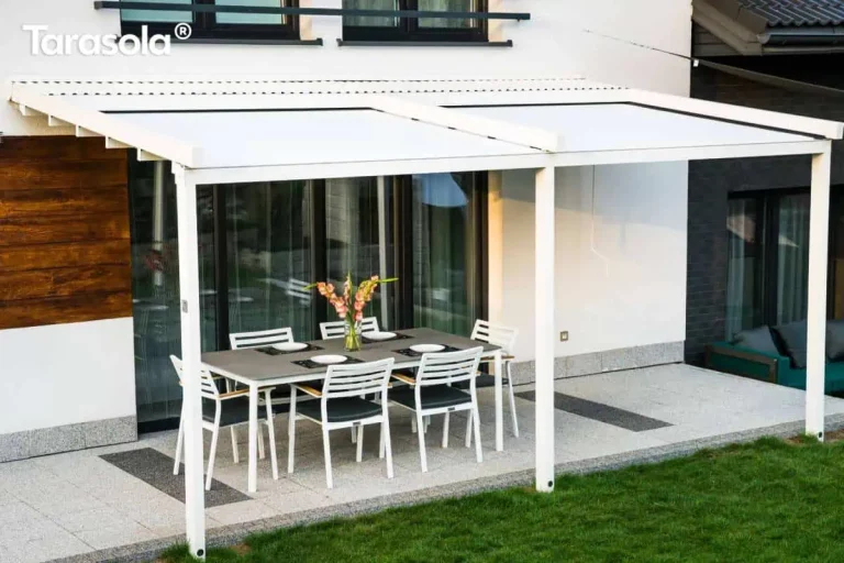Szeroka, biała pergola tarasowa Tarasola Simple o konstrukcji przyściennej, zamontowana na elewacji nowoczesnego domu jednorodzinnego. Zadaszenie składa się z dwóch sekcji białego, napiętego materiału technicznego, zamocowanego na aluminiowych profilach poprzecznych i prowadnicach. Konstrukcja opiera się na trzech pionowych słupach o kwadratowym przekroju, które są zakotwiczone w gruncie, na trawniku przy krawędzi tarasu. Pod zadaszeniem znajduje się strefa wypoczynkowa z dużym, prostokątnym stołem o szarym blacie oraz sześcioma białymi krzesłami z ciemnymi poduszkami. Taras został wyłożony jasnymi, nakrapianymi płytami z ozdobnymi ciemniejszymi pasami. Elewacja budynku łączy biały tynk z panelami imitującymi naturalne drewno w ciepłym brązowym odcieniu. W tle widoczne są duże przeszklenia drzwi tarasowych, fragment zielonego trawnika oraz sąsiednia zabudowa. Zdjęcie wykonane z perspektywy bocznej, ukazujące znaczną szerokość konstrukcji i jej integrację z bryłą budynku.