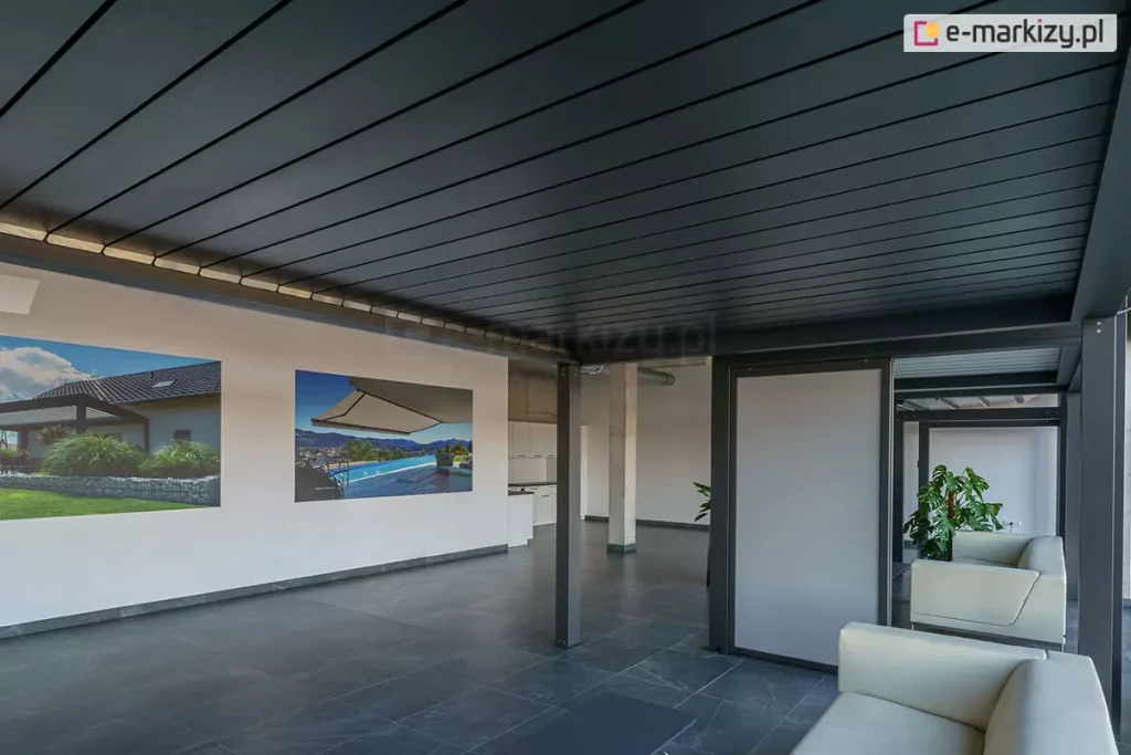 Widok od wewnątrz na system zadaszenia pergoli tarasowej Sb350 Aluprof w kolorze antracytowym z całkowicie zamkniętymi lamelami. Lamele tworzą jednolitą, szczelną płaszczyznę dachu. Pod pergolą znajduje się salon ekspozycyjny z ciemnoszarą podłogą, na której ustawiono jasne meble wypoczynkowe (sofy). Na białej ścianie w tle zawieszone są dwa duże plakaty przedstawiające realizacje pergoli i markiz w ogrodach. Po prawej stronie widoczny jest pionowy panel przesuwny (screen) oraz fragment kolejnej strefy ekspozycyjnej z roślinnością i betonowym filarem.