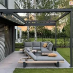 Nowoczesny taras ogrodowy z prostą, elegancką pergolą wykonaną z ciemnoszarego metalu. Konstrukcja ma prostokątną ramę dachową z poprzecznymi belkami, ze zwiniętą do ściany nad tarasem tkaniną dachu, której belki przeciwwietrzne wyposażono we wbudowane punkty świetlne, które są włączone. Pod pergolą ustawiono nowoczesny zestaw wypoczynkowy – modułową sofę narożną i leżankę z tej samej serii, na drewnianych stelażach z metalowymi nogami. Meble są tapicerowane w kolorze jasnoszarym, z dekoracyjnymi poduszkami w odcieniach żółci i szarości. Pomiędzy elementami ustawiono niski stolik kawowy Taras wyłożony jest dużymi płytami w odcieniu jasnej szarości. Otoczenie stanowi wypielęgnowany trawnik, żywopłot i ogrodzenie z siatki. Za ogrodzeniem rosną drzewa liściaste, w tym kilka brzóz z białą korą. W tle widoczna jest sąsiednia zabudowa. Po lewej stronie kadru widać fragment domu z dużym oknem osłoniętym poziomymi, zewnętrznymi żaluzjami w ciemnym kolorze. Całość tworzy uporządkowaną, nowoczesną przestrzeń wypoczynkową.