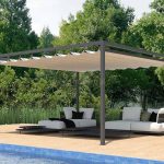 Wizualizacja wolnostojącej pergoli ogrodowej o minimalistycznej, antracytowej konstrukcji aluminiowej, ustawionej na jasnym, drewnianym tarasie przy basenie. Zadaszenie wykonane z jasnobeżowej tkaniny akrylowej jest rozłożone, tworząc regularne, dekoracyjne pofalowania między poprzecznymi profilami prowadzącymi. Pod pergolą zaaranżowano nowoczesną strefę wypoczynkową z białymi, niskimi modułowymi sofami ozdobionymi ciemnymi poduszkami. Meble spoczywają na płaskich, czarnych platformach, które pełnią funkcję zintegrowanych stolików bocznych. W dolnej części kadru widoczny jest fragment basenu z błękitną mozaiką oraz dwa brązowe, ergonomiczne leżaki ustawione na brzegu tarasu. Tło stanowi gęsta ściana wysokich, zielonych drzew oraz pas niskich traw ozdobnych, które oddzielają strefę tarasu od naturalnego krajobrazu. Całość ujęcia prezentuje się w pełnym, dziennym świetle, podkreślając luksusowy charakter letniej strefy relaksu.