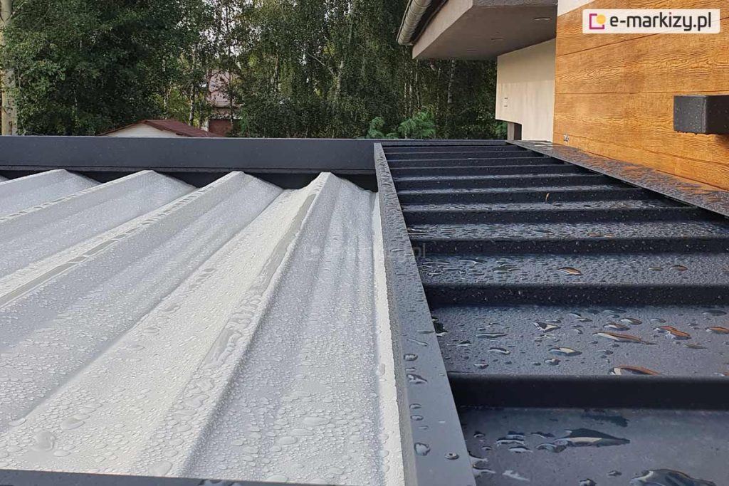 Zdjęcie prezentuje zbliżenie na górną część pergoli tarasowej Luxury podczas opadów deszczu, ukazując system ochrony poszycia po jego złożeniu oraz wodoodporność tkaniny. Po lewej stronie widoczne jest jasne, teksturowane poszycie dachowe, na którym osadziły się liczne krople wody. Po prawej stronie znajduje się stały daszek ochronny wykonany z blachy lub profilu aluminiowego w kolorze antracytowym, również pokryty kroplami deszczu, który osłania zwinięty materiał przed czynnikami atmosferycznymi i zabrudzeniami. Konstrukcja daszka posiada poprzeczne przetłoczenia wzmacniające i jest zamontowana bezpośrednio przy elewacji budynku o strukturze imitującej drewno. W tle widoczne są korony drzew oraz fragment dachu sąsiedniego budynku, co podkreśla skuteczność systemu odprowadzania wody i ochrony tkaniny w warunkach zewnętrznych.