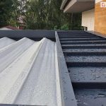 Zdjęcie prezentuje zbliżenie na górną część pergoli tarasowej Luxury podczas opadów deszczu, ukazując system ochrony poszycia po jego złożeniu oraz wodoodporność tkaniny. Po lewej stronie widoczne jest jasne, teksturowane poszycie dachowe, na którym osadziły się liczne krople wody. Po prawej stronie znajduje się stały daszek ochronny wykonany z blachy lub profilu aluminiowego w kolorze antracytowym, również pokryty kroplami deszczu, który osłania zwinięty materiał przed czynnikami atmosferycznymi i zabrudzeniami. Konstrukcja daszka posiada poprzeczne przetłoczenia wzmacniające i jest zamontowana bezpośrednio przy elewacji budynku o strukturze imitującej drewno. W tle widoczne są korony drzew oraz fragment dachu sąsiedniego budynku, co podkreśla skuteczność systemu odprowadzania wody i ochrony tkaniny w warunkach zewnętrznych.