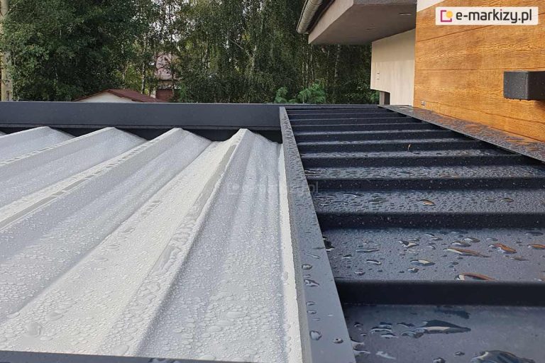 Zdjęcie prezentuje zbliżenie na górną część pergoli tarasowej Luxury podczas opadów deszczu, ukazując system ochrony poszycia po jego złożeniu oraz wodoodporność tkaniny. Po lewej stronie widoczne jest jasne, teksturowane poszycie dachowe, na którym osadziły się liczne krople wody. Po prawej stronie znajduje się stały daszek ochronny wykonany z blachy lub profilu aluminiowego w kolorze antracytowym, również pokryty kroplami deszczu, który osłania zwinięty materiał przed czynnikami atmosferycznymi i zabrudzeniami. Konstrukcja daszka posiada poprzeczne przetłoczenia wzmacniające i jest zamontowana bezpośrednio przy elewacji budynku o strukturze imitującej drewno. W tle widoczne są korony drzew oraz fragment dachu sąsiedniego budynku, co podkreśla skuteczność systemu odprowadzania wody i ochrony tkaniny w warunkach zewnętrznych.