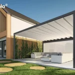 Wizualizacja przedstawia nowoczesną pergolę tarasową Tarasola Flat o konstrukcji wolnostojącej, zainstalowaną na przestronnym tarasie z jasnych, kompozytowych desek. Aluminiowa rama w kolorze głębokiego antracytu wspiera białe, tkaninowe zadaszenie rzymskie, które jest rozłożone i układa się w charakterystyczne, miękkie fale podwieszone na poprzecznych profilach. Konstrukcja charakteryzuje się masywną górną belką, która pełni funkcję estetyczną oraz techniczną, skrywając system odprowadzania wody. Pod pergolą zaaranżowano luksusową strefę wypoczynku z białym meblem narożnym i niskim stolikiem kawowym, osłoniętą w pełni rozwiniętym refleksolem Zip. W tle widoczny jest nowoczesny budynek z elewacją łączącą biel, cegłę oraz duże przeszklenia, a także starannie zaprojektowany ogród z trawnikiem i ozdobnymi trawami.
