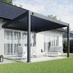 Pergola Rubio Soft z bioklimatycznym dachem lamelowym sterowanym ręczną korbą, z aluminiowymi lamelami ustawionymi w pozycji półotwartej, tworzącymi charakterystyczny cień na tarasie. Konstrukcja w grafitowym kolorze przylega do elewacji budynku, a pod nią znajduje się jasny zestaw wypoczynkowy ustawiony na tarasie otoczonym trawnikiem.