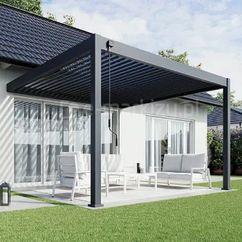 Pergola Rubio Soft z bioklimatycznym dachem lamelowym sterowanym ręczną korbą, z aluminiowymi lamelami ustawionymi w pozycji półotwartej, tworzącymi charakterystyczny cień na tarasie. Konstrukcja w grafitowym kolorze przylega do elewacji budynku, a pod nią znajduje się jasny zestaw wypoczynkowy ustawiony na tarasie otoczonym trawnikiem.