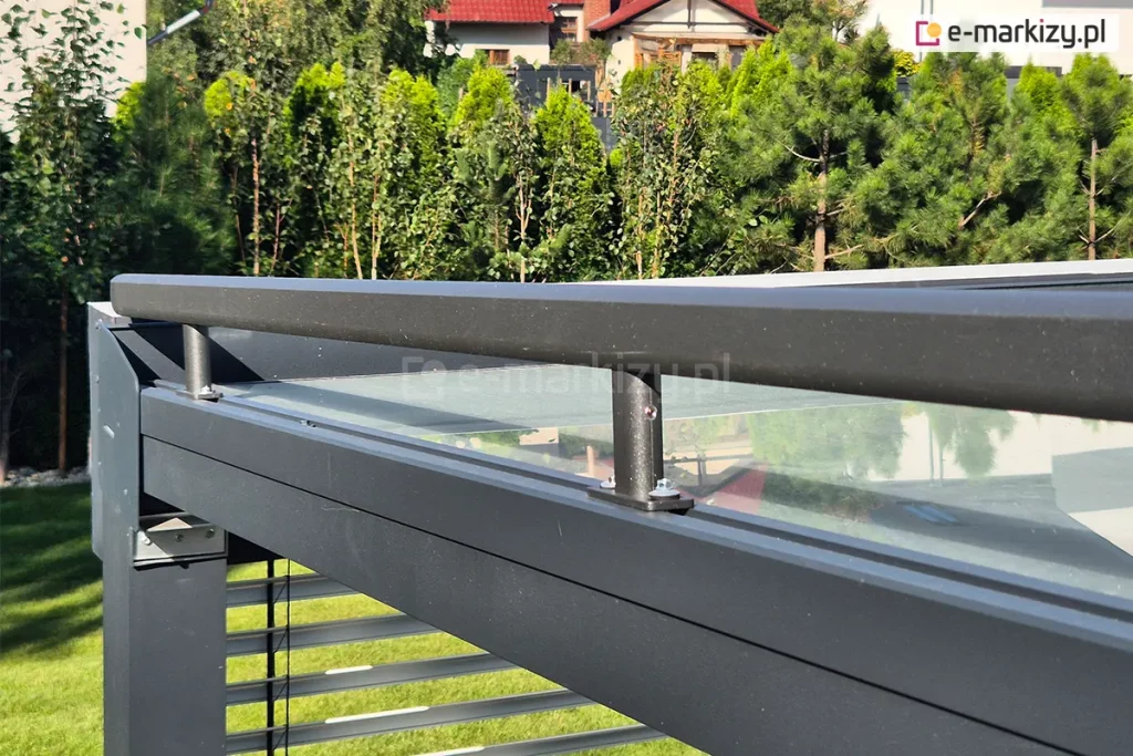 Widok z boku na konstrukcję nośną pergoli tarasowej z zainstalowanym systemem osłonowym na dachu - roleta veranda. Na pierwszym planie widoczny jest masywny, poziomy profil aluminiowy w kolorze antracytowym, do którego przytwierdzona jest szyna prowadząca wsparta na pionowych uchwytach. Całość zamontowana jest nad szklanym zadaszeniem, w którym odbija się otoczenie. W lewej części kadru widoczny jest słupek konstrukcyjny oraz fragment żaluzji fasadowej (lameli) osłaniającej bok pergoli. W tle znajduje się zadbany ogród z gęstym żywopłotem z tui, krzewami oraz fragmentami dachów domów jednorodzinnych. Zdjęcie prezentuje stabilność i precyzyjne łączenie elementów nowoczesnej zabudowy tarasu.