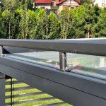 Widok z boku na konstrukcję nośną pergoli tarasowej z zainstalowanym systemem osłonowym na dachu - roleta veranda. Na pierwszym planie widoczny jest masywny, poziomy profil aluminiowy w kolorze antracytowym, do którego przytwierdzona jest szyna prowadząca wsparta na pionowych uchwytach. Całość zamontowana jest nad szklanym zadaszeniem, w którym odbija się otoczenie. W lewej części kadru widoczny jest słupek konstrukcyjny oraz fragment żaluzji fasadowej (lameli) osłaniającej bok pergoli. W tle znajduje się zadbany ogród z gęstym żywopłotem z tui, krzewami oraz fragmentami dachów domów jednorodzinnych. Zdjęcie prezentuje stabilność i precyzyjne łączenie elementów nowoczesnej zabudowy tarasu.