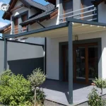 Nowoczesna pergola tarasowa Simple marki Tarasola o konstrukcji przyściennej w kolorze ciemnoszarym (antracytowym), zamontowana nad tarasem wyłożonym dużymi, grafitowymi płytami. Zadaszenie stanowi ruchoma tkanina techniczna w kolorze kremowym, która jest rozwinięta, odsłaniając widok na aluminiowe profile prowadzące. Konstrukcja wspiera się na dwóch solidnych słupach o przekroju kwadratowym, a jedna z bocznych stron została wyposażona w pełny ekran osłonowy w kolorze dopasowanym do stelaża, zapewniający prywatność i ochronę przed wiatrem. Budynek o jasnej elewacji posiada ciemne obramowania wokół okien tarasowych oraz dopasowane kolorystycznie barierki balkonowe na piętrze. Na tarasie oraz w jego bezpośrednim sąsiedztwie widoczne są rośliny ozdobne w donicach i nasadzenia ogrodowe, w tym niskie krzewy i drzewka formowane w kule. Zdjęcie wykonane w pełnym słońcu, podkreślającym kontrast między jasnym materiałem dachu a ciemną konstrukcją pergoli.