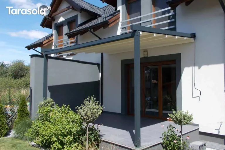 Nowoczesna pergola tarasowa Simple marki Tarasola o konstrukcji przyściennej w kolorze ciemnoszarym (antracytowym), zamontowana nad tarasem wyłożonym dużymi, grafitowymi płytami. Zadaszenie stanowi ruchoma tkanina techniczna w kolorze kremowym, która jest rozwinięta, odsłaniając widok na aluminiowe profile prowadzące. Konstrukcja wspiera się na dwóch solidnych słupach o przekroju kwadratowym, a jedna z bocznych stron została wyposażona w pełny ekran osłonowy w kolorze dopasowanym do stelaża, zapewniający prywatność i ochronę przed wiatrem. Budynek o jasnej elewacji posiada ciemne obramowania wokół okien tarasowych oraz dopasowane kolorystycznie barierki balkonowe na piętrze. Na tarasie oraz w jego bezpośrednim sąsiedztwie widoczne są rośliny ozdobne w donicach i nasadzenia ogrodowe, w tym niskie krzewy i drzewka formowane w kule. Zdjęcie wykonane w pełnym słońcu, podkreślającym kontrast między jasnym materiałem dachu a ciemną konstrukcją pergoli.