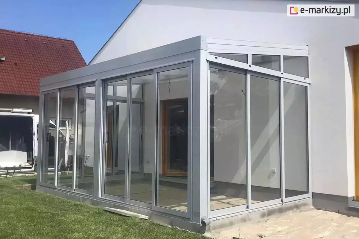 Nowoczesna, biała pergola tarasowa Modes, która została w pełni zabudowana szklanymi panelami, tworząc estetyczny ogród zimowy. Konstrukcja przylega do białej elewacji domu i posiada jednospadowy dach z wyraźnymi profilami. Frontową oraz boczną ścianę tworzą przesuwne, transparentne tafle szkła osadzone w aluminiowych ramach, przy czym boczne przeszklenia pod skosem dachu (tzw. kliny) są zamontowane na stałe. Wewnątrz, na podłożu z jasnych płyt, widoczne są drewniane drzwi prowadzące do budynku, a cała konstrukcja osadzona jest na niskim fundamencie przy zielonym trawniku. W tle, pod bezchmurnym niebem, widać fragment białego pojazdu serwisowego oraz sąsiedni dom z dachem z czerwonej dachówki.