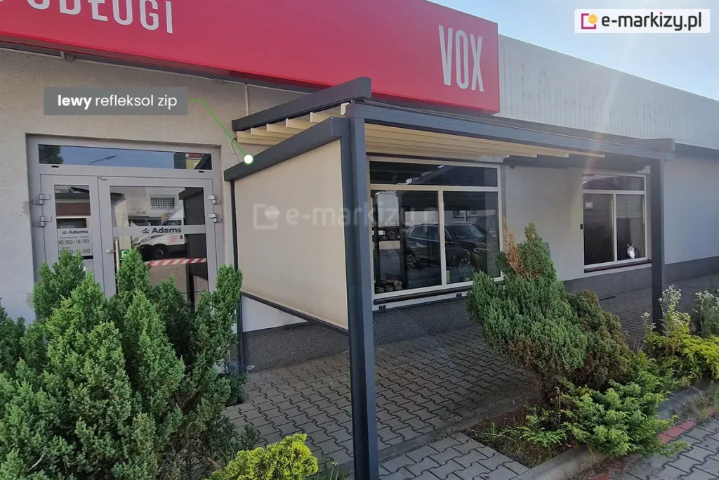 Zdjęcie przedstawia nowoczesną pergolę tarasową zainstalowaną przy niskim, komercyjnym budynku z szarą elewacją i dużymi witrynami. Konstrukcja pergoli jest wykonana z grubych profili aluminiowych w kolorze ciemnego antracytu i wspiera się na pionowych słupach o kwadratowym przekroju. Zadaszenie stanowi jasnokremowa tkanina ułożona w regularne fałdy, zamontowana pod lekkim skosem. Na lewym boku konstrukcji zainstalowano pionową osłonę zewnętrzną, którą wskazuje zielona linia pomocnicza z podpisem w szarej ramce: „lewy refleksol zip”. Tkanina refleksola jest w kolorze beżowym, półtransparentna i całkowicie rozwinięta, zasłaniając boczną przestrzeń tarasu. Podłoże przed budynkiem wyłożone jest szarą kostką brukową typu behaton, a wzdłuż elewacji rosną ozdobne, gęste krzewy iglaste. Nad wejściem do budynku widoczny jest fragment czerwonego szyldu z białym napisem oraz logotyp marki VOX. Całość ujęcia wykonano w świetle dziennym, prezentując kompletny system osłonowy integrujący zadaszenie poziome z boczną roletą pionową.