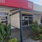 Zdjęcie przedstawia nowoczesną pergolę tarasową zainstalowaną przy niskim, komercyjnym budynku z szarą elewacją i dużymi witrynami. Konstrukcja pergoli jest wykonana z grubych profili aluminiowych w kolorze ciemnego antracytu i wspiera się na pionowych słupach o kwadratowym przekroju. Zadaszenie stanowi jasnokremowa tkanina ułożona w regularne fałdy, zamontowana pod lekkim skosem. Na lewym boku konstrukcji zainstalowano pionową osłonę zewnętrzną, którą wskazuje zielona linia pomocnicza z podpisem w szarej ramce: „lewy refleksol zip”. Tkanina refleksola jest w kolorze beżowym, półtransparentna i całkowicie rozwinięta, zasłaniając boczną przestrzeń tarasu. Podłoże przed budynkiem wyłożone jest szarą kostką brukową typu behaton, a wzdłuż elewacji rosną ozdobne, gęste krzewy iglaste. Nad wejściem do budynku widoczny jest fragment czerwonego szyldu z białym napisem oraz logotyp marki VOX. Całość ujęcia wykonano w świetle dziennym, prezentując kompletny system osłonowy integrujący zadaszenie poziome z boczną roletą pionową.