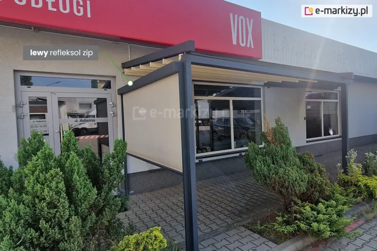 Zdjęcie przedstawia nowoczesną pergolę tarasową zainstalowaną przy niskim, komercyjnym budynku z szarą elewacją i dużymi witrynami. Konstrukcja pergoli jest wykonana z grubych profili aluminiowych w kolorze ciemnego antracytu i wspiera się na pionowych słupach o kwadratowym przekroju. Zadaszenie stanowi jasnokremowa tkanina ułożona w regularne fałdy, zamontowana pod lekkim skosem. Na lewym boku konstrukcji zainstalowano pionową osłonę zewnętrzną, którą wskazuje zielona linia pomocnicza z podpisem w szarej ramce: „lewy refleksol zip”. Tkanina refleksola jest w kolorze beżowym, półtransparentna i całkowicie rozwinięta, zasłaniając boczną przestrzeń tarasu. Podłoże przed budynkiem wyłożone jest szarą kostką brukową typu behaton, a wzdłuż elewacji rosną ozdobne, gęste krzewy iglaste. Nad wejściem do budynku widoczny jest fragment czerwonego szyldu z białym napisem oraz logotyp marki VOX. Całość ujęcia wykonano w świetle dziennym, prezentując kompletny system osłonowy integrujący zadaszenie poziome z boczną roletą pionową.
