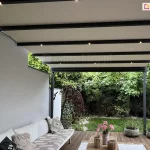 Zdjęcie prezentuje perspektywę spod pergoli tarasowej Aura, ukazującą detale jej zadaszenia oraz aranżację strefy wypoczynkowej. Konstrukcja pergoli opiera się na aluminiowych profilach w kolorze głębokiego antracytu, a jej dach tworzy jasna, kremowa tkanina rozpięta na poprzecznych belkach. W co drugą z belek wbudowano punktowe oświetlenie LED, które emituje ciepłe, złociste światło, oświetlając przestrzeń tarasu po zmroku. Lewa strona konstrukcji przylega bezpośrednio do gładkiej, jasnoszarej ściany budynku. Pod zadaszeniem widoczny jest fragment narożnej sofy ogrodowej z jasnymi siedziskami, na której ułożone są dekoracyjne poduszki – jedna czarna z napisem oraz beżowa z ozdobnymi frędzlami i geometrycznym wzorem. Podłoga tarasu wykonana jest z ciemnobrązowych desek o widocznej fakturze drewna. Na drewnianym stoliku kawowym, widocznym w dolnej części kadru, ustawiono wazon z różowymi kwiatami oraz dwa dekoracyjne, plecione lampiony. W tle rozpościera się widok na gęsty, zielony ogród. Bezpośrednio przy tarasie rośnie ozdobne drzewko o ciemnoczerwonych, purpurowych liściach, a obok słupa konstrukcyjnego ustawiono dwie okrągłe, betonowe donice z kępami ozdobnych traw. Dalszy plan wypełnia soczysta zieleń trawnika oraz ściana liściastych drzew i krzewów, które zapewniają domownikom prywatność i naturalne tło dla nowoczesnej architektury tarasu.