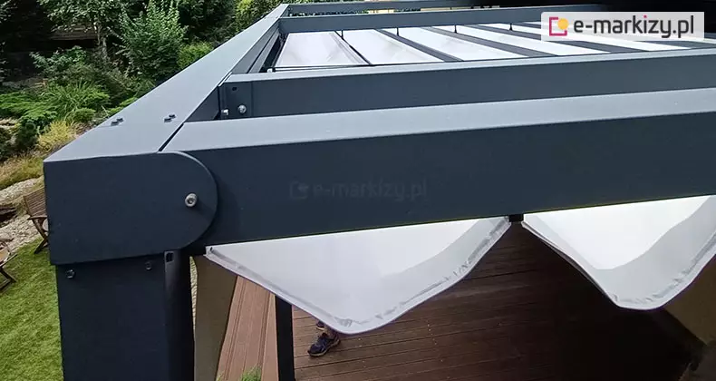 Zbliżenie na górny narożnik i system zadaszenia pergoli rzymskiej Tako w kolorze antracytowym. Konstrukcja wykonana jest z grubych profili aluminiowych o matowym wykończeniu, łączonych za pomocą widocznych śrub. Pod ramą dachu zamontowane jest segmentowe poszycie z białej tkaniny technicznej, która układa się w dekoracyjne, luźne fale (fałdy). Tkanina podwieszona jest na poprzecznych profilach prowadzących. W tle, pod zadaszeniem, widoczny jest taras z desek drewnianych, a na zewnątrz zielony ogród z trawnikiem i gęstymi krzewami. Perspektywa zdjęcia z góry ukazuje detale montażowe narożnika oraz sposób mocowania materiału osłonowego.