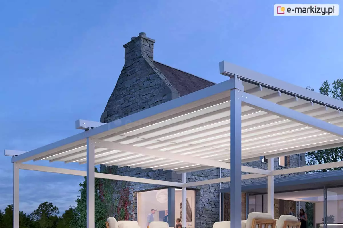 Pergola wolnostojąca Solid Aluprof z podświetlonym dachem tkaninowym ustawiona na tarasie w pobliżu domu wykonanego z kamienia. W budynku, przez duże okna, widoczni ludzie. Nad tarasem widoczne ciemniejące niebo.