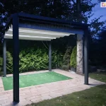 Antracytowa pergola Tako wolnostojąca z białym dachem rozwiniętym na całości konstrukcji oraz włączonym oświetleniem led w belkach przeciwwietrznych. Jasne światło pergoli oświetla patio ogrodowe wyłożone kostką przy trawniku, na którym leży kawałek zielonej imitującej trawę tkaniny oraz rośliny rosnące w pobliżu miejsca z pergolą.