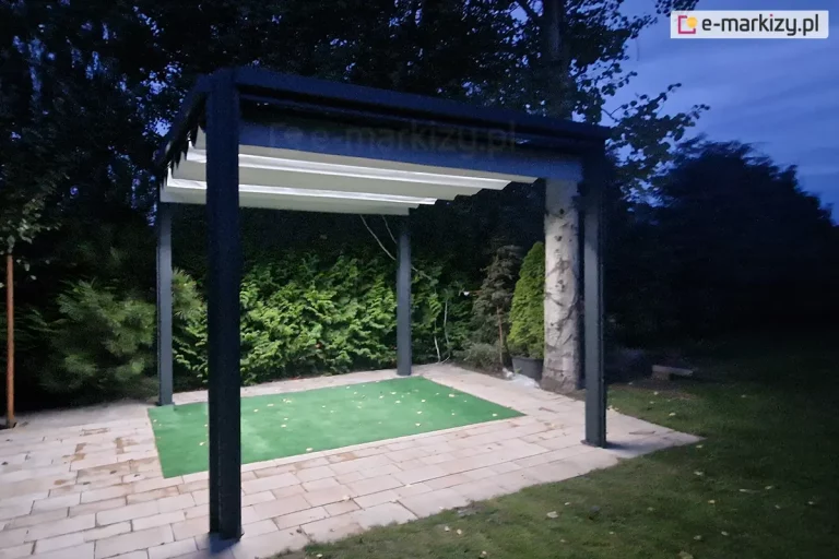 Antracytowa pergola Tako wolnostojąca z białym dachem rozwiniętym na całości konstrukcji oraz włączonym oświetleniem led w belkach przeciwwietrznych. Jasne światło pergoli oświetla patio ogrodowe wyłożone kostką przy trawniku, na którym leży kawałek zielonej imitującej trawę tkaniny oraz rośliny rosnące w pobliżu miejsca z pergolą.
