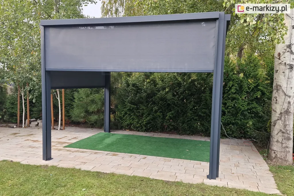 Frontowy widok na aluminiową pergolę tarasową w kolorze ciemnego antracytu. Przednia roleta typu Screen jest opuszczona do około 1/3 wysokości, prezentując strukturę drobnej siatki technicznej. Boczne rolety są opuszczone do połowy, tworząc osłoniętą przestrzeń wypoczynkową. Konstrukcja wsparta na czterech pionowych słupach znajduje się na utwardzonym tarasie z kostki. W tle widać gęste zielone krzewy iglaste oraz pnie drzew, które stanowią naturalne tło dla antracytowej ramy konstrukcji.