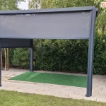 Frontowy widok na aluminiową pergolę tarasową w kolorze ciemnego antracytu. Przednia roleta typu Screen jest opuszczona do około 1/3 wysokości, prezentując strukturę drobnej siatki technicznej. Boczne rolety są opuszczone do połowy, tworząc osłoniętą przestrzeń wypoczynkową. Konstrukcja wsparta na czterech pionowych słupach znajduje się na utwardzonym tarasie z kostki. W tle widać gęste zielone krzewy iglaste oraz pnie drzew, które stanowią naturalne tło dla antracytowej ramy konstrukcji.
