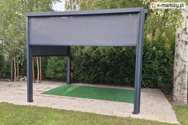 Frontowy widok na aluminiową pergolę tarasową w kolorze ciemnego antracytu. Przednia roleta typu Screen jest opuszczona do około 1/3 wysokości, prezentując strukturę drobnej siatki technicznej. Boczne rolety są opuszczone do połowy, tworząc osłoniętą przestrzeń wypoczynkową. Konstrukcja wsparta na czterech pionowych słupach znajduje się na utwardzonym tarasie z kostki. W tle widać gęste zielone krzewy iglaste oraz pnie drzew, które stanowią naturalne tło dla antracytowej ramy konstrukcji.