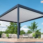 Pergola lamelowa wolnostojąca Bioline, bioklimatyczna o nowoczesnej, geometrycznej konstrukcji, ustawiona na podwyższonym tarasie przy basenie. Aluminiowa konstrukcja pergoli w kolorze głębokiego antracytu składa się z grubych profili ramowych oraz czterech masywnych słupów nośnych o kwadratowym przekroju. Zadaszenie tworzy system poziomych, białych lameli, które są ustawione pod kątem, pozwalając na regulację dopływu światła słonecznego. Pod pergolą znajduje się luksusowa strefa wypoczynkowa wyposażona w meble ogrodowe z technorattanu o jasnej plecionce i szarych poduchach, w tym sofy oraz stolik kawowy. Po bokach mebli ustawiono symetrycznie donice z zielonymi roślinami o podłużnych liściach. Taras wykonany jest z płyt o teksturze jasnego kamienia lub betonu i sąsiaduje bezpośrednio z krawędzią basenu z czystą, błękitną wodą. W tle widoczna jest gęsta ściana zieleni złożona z drzew i krzewów liściastych oraz czarne, nowoczesne ogrodzenie panelowe. Całość kompozycji dopełnia bezchmurne, jasnoniebieskie niebo z delikatnymi, białymi obłokami, co podkreśla letni i wypoczynkowy charakter prezentowanej architektury ogrodowej.