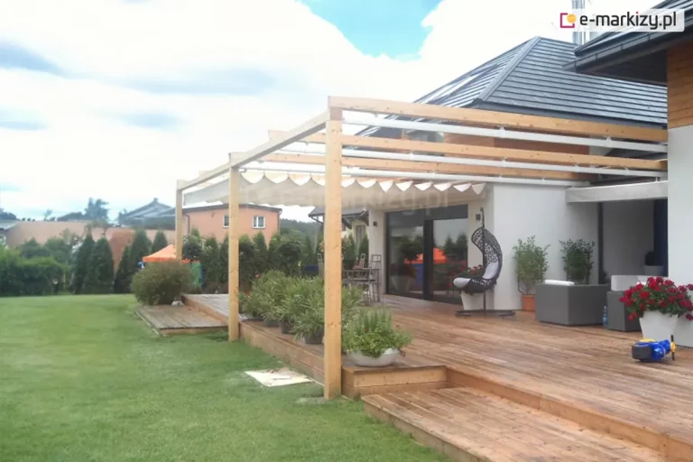 Zdjęcie prezentuje szeroką perspektywę na duży taras wykonany z naturalnego drewna, nad którym zamontowano pergolę z systemem rozsuwanego dachu Silva. Konstrukcja pergoli opiera się na jasnych, drewnianych belkach i słupach, tworzących otwarty szkielet nad przestrzenią wypoczynkową. Jasne, kremowe poszycie dachu jest częściowo zsunięte, ukazując segmentową strukturę materiału ułożonego w równe fale. Taras jest wielopoziomowy, wyposażony w schody prowadzące na zadbany, zielony trawnik otoczony żywopłotem z tui. Na deskach tarasowych ustawiono liczne donice z roślinnością, nowoczesne meble ogrodowe w kolorze szarym oraz wiszący fotel typu kokon. W tle widoczna jest jasna elewacja domu jednorodzinnego z dużymi przeszkleniami oraz sąsiednie budynki pod zachmurzonym niebem. Cała aranżacja podkreśla nowoczesny i komfortowy charakter strefy relaksu na świeżym powietrzu.