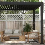 Nowoczesna pergola tarasowa w kolorze antracytowym z otwartym dachem lamelowym, zamontowana na szarym podeście przy jasnej, poziomej elewacji budynku. Konstrukcja opiera się na czterech ciemnych, matowych profilach aluminiowych, a dach tworzą równolegle ustawione czarne lamele. Wewnątrz zaaranżowano strefę wypoczynkową z drewnianą sofą i dwoma fotelami o jasnej tapicerce. Pod meblami znajduje się wzorzysty dywan zewnętrzny oraz okrągła, pleciona mata. Dodatki stanowią plecione pufy, stoliki pomocnicze z drewna i wikliny oraz liczne rośliny doniczkowe, w tym palmy i kwiaty w białych donicach. Tło pergoli stanowi białe ogrodzenie z ozdobną kratownicą w górnej części. Z lewej strony górnej ramy zwisa metalowy drążek korbowy służący do sterowania zadaszeniem.