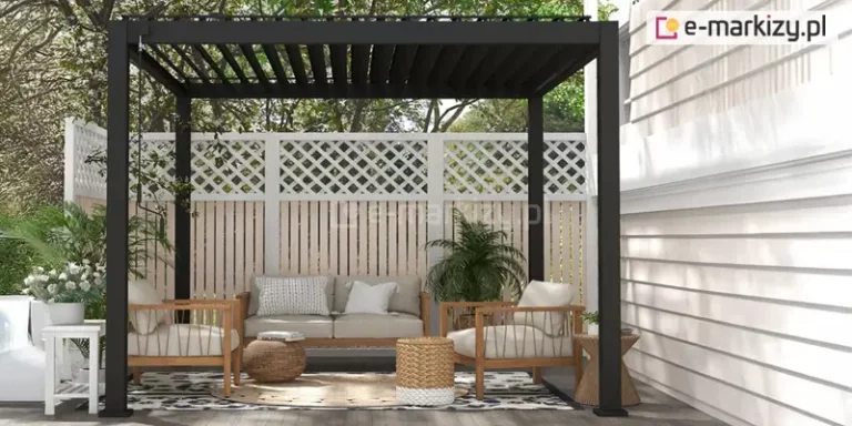 Nowoczesna pergola lamelowa wolnostojąca Gloria w kolorze antracytowym z otwartym dachem lamelowym, zamontowana na szarym podeście przy jasnej, poziomej elewacji budynku. Konstrukcja opiera się na czterech ciemnych, matowych profilach aluminiowych, a dach tworzą równolegle ustawione czarne lamele. Wewnątrz zaaranżowano strefę wypoczynkową z drewnianą sofą i dwoma fotelami o jasnej tapicerce. Pod meblami znajduje się wzorzysty dywan zewnętrzny oraz okrągła, pleciona mata. Dodatki stanowią plecione pufy, stoliki pomocnicze z drewna i wikliny oraz liczne rośliny doniczkowe, w tym palmy i kwiaty w białych donicach. Tło pergoli stanowi białe ogrodzenie z ozdobną kratownicą w górnej części. Z lewej strony górnej ramy zwisa metalowy drążek korbowy służący do sterowania zadaszeniem.