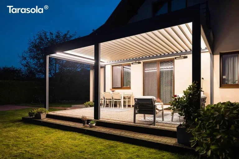 Zdjęcie przedstawia nowoczesną, bioklimatyczną konstrukcję Essential tarasola w warunkach nocnych, zamontowaną przy jasnej elewacji domu. Pergola w kolorze ciemnego antracytu jest intensywnie oświetlona od wewnątrz za pomocą zintegrowanych listew LED, które emitują jasne, ciepłe światło, oświetlając białe lamele dachowe oraz przestrzeń tarasu. Pod zadaszeniem widoczna jest strefa jadalniana z białym stołem i krzesłami oraz strefa wypoczynkowa z wygodnym fotelem, ustawione na dekoracyjnych dywanach zewnętrznych. Na ścianie budynku, pod dachem pergoli, zamontowane są dwa promienniki ciepła, które zwiększają komfort użytkowania tarasu w chłodniejsze wieczory. Konstrukcja wsparta jest na solidnych słupach, a podłoże tarasu wyłożono jasną kostką brukową z ciemniejszym obramowaniem. W tle, za oświetloną strefą, panuje głęboki mrok nocy, z ledwo widocznymi zarysami drzew i ogrodu, co potęguje kontrast i skupia uwagę na przytulnie urządzonej przestrzeni pod pergolą.