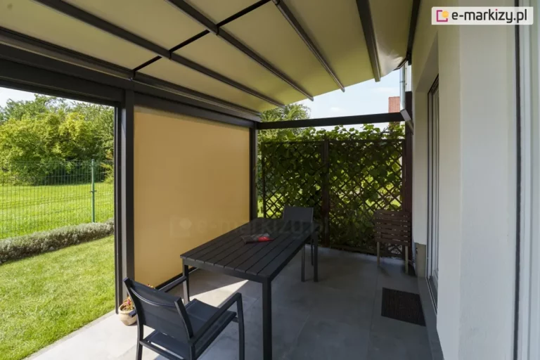 Widok od wewnątrz na nowoczesną pergolę tarasową Solid o ciemnej, antracytowej konstrukcji aluminiowej, zamontowaną przy jasnej elewacji domu. Zadaszenie stanowi ruchoma, segmentowa tkanina w kolorze kremowym, rozpięta na poprzecznych profilach. Jeden z bocznych segmentów pergoli został wyposażony w pionową osłonę przeciwsłoneczną typu refleksol w kolorze żółtym, która zapewnia prywatność i ochronę przed słońcem. Na tarasie wyłożonym szarymi płytami gresowymi znajduje się strefa jadalniana z czarnym, prostokątnym stołem oraz dopasowanymi kolorystycznie nowoczesnymi krzesłami z podłokietnikami. W tle widoczna jest ażurowa drewniana kratka porośnięta gęstą roślinnością pnącą, a po lewej stronie kadru rozpościera się widok na zielony trawnik ogrodzony niskim żywopłotem i siatką. Zdjęcie wykonane w pełnym świetle dziennym, podkreślające funkcjonalność osłon bocznych i systemów zadaszeń tkaninowych.