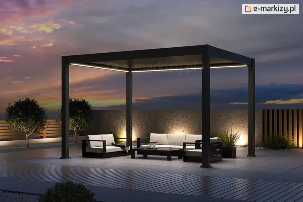 Wolnostojąca pergola bioklimatyczna Rubio w kolorze antracytowym, zaprezentowana w aranżacji wieczornej z włączonym systemem oświetlenia LED. Liniowe paski LED o ciepłej barwie, zamontowane wzdłuż wewnętrznych krawędzi górnej ramy, oświetlają strefę wypoczynkową, na którą składają się nowoczesne meble ogrodowe z jasnymi poduszkami. Konstrukcja z dachem lamelowym w pozycji zamkniętej posadowiona jest na tarasie z jasnych płyt betonowych, sąsiadującym z nawierzchnią z ciemnej deski kompozytowej. Tło tworzy surowy, betonowy mur z punktowym podświetleniem dolnym, a po prawej stronie widoczne jest ażurowe ogrodzenie z pionowych listew. Scenerię dopełniają dwa formowane drzewka w pasie białego grysu oraz rozległy widok na niebo w odcieniach fioletu i pomarańczu podczas zachodu słońca. Całość prezentuje estetykę produktu w warunkach nocnych, podkreślając funkcjonalność zintegrowanego oświetlenia.