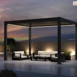 Wolnostojąca pergola bioklimatyczna Rubio w kolorze antracytowym, zaprezentowana w aranżacji wieczornej z włączonym systemem oświetlenia LED. Liniowe paski LED o ciepłej barwie, zamontowane wzdłuż wewnętrznych krawędzi górnej ramy, oświetlają strefę wypoczynkową, na którą składają się nowoczesne meble ogrodowe z jasnymi poduszkami. Konstrukcja z dachem lamelowym w pozycji zamkniętej posadowiona jest na tarasie z jasnych płyt betonowych, sąsiadującym z nawierzchnią z ciemnej deski kompozytowej. Tło tworzy surowy, betonowy mur z punktowym podświetleniem dolnym, a po prawej stronie widoczne jest ażurowe ogrodzenie z pionowych listew. Scenerię dopełniają dwa formowane drzewka w pasie białego grysu oraz rozległy widok na niebo w odcieniach fioletu i pomarańczu podczas zachodu słońca. Całość prezentuje estetykę produktu w warunkach nocnych, podkreślając funkcjonalność zintegrowanego oświetlenia.