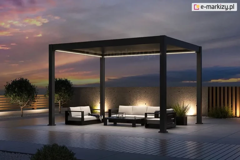 Wolnostojąca pergola bioklimatyczna Rubio w kolorze antracytowym, zaprezentowana w aranżacji wieczornej z włączonym systemem oświetlenia LED. Liniowe paski LED o ciepłej barwie, zamontowane wzdłuż wewnętrznych krawędzi górnej ramy, oświetlają strefę wypoczynkową, na którą składają się nowoczesne meble ogrodowe z jasnymi poduszkami. Konstrukcja z dachem lamelowym w pozycji zamkniętej posadowiona jest na tarasie z jasnych płyt betonowych, sąsiadującym z nawierzchnią z ciemnej deski kompozytowej. Tło tworzy surowy, betonowy mur z punktowym podświetleniem dolnym, a po prawej stronie widoczne jest ażurowe ogrodzenie z pionowych listew. Scenerię dopełniają dwa formowane drzewka w pasie białego grysu oraz rozległy widok na niebo w odcieniach fioletu i pomarańczu podczas zachodu słońca. Całość prezentuje estetykę produktu w warunkach nocnych, podkreślając funkcjonalność zintegrowanego oświetlenia.