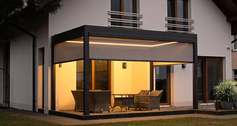 Nowoczesna pergola tarasowa w kolorze antracytowym, zamontowana przy białej elewacji piętrowego domu, sfotografowana wieczorem przy włączonym oświetleniu. Konstrukcja posiada zintegrowane oświetlenie LED w formie liniowej listwy biegnącej wzdłuż wewnętrznej ramy dachu, oświetlającej wnętrze ciepłym światłem. Dwa boki pergoli są osłonięte półtransparentnymi roletami bocznymi typu screen w kolorze jasnoszarym, które są opuszczone do połowy wysokości. Pod zadaszeniem znajduje się zestaw mebli ogrodowych z technorattanu, składający się z foteli i stolika. Na piętrze budynku widoczne są dwa okna z brązowymi ramami i białymi barierkami. Po prawej stronie tarasu, na zewnątrz pergoli, stoją ozdobne donice z kępami traw, a w tle widać ciemne sylwetki drzew na tle wieczornego nieba.