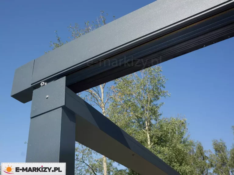 Zbliżenie na łączenie profili aluminiowych w kolorze antracytowym stanowiących solidną konstrukcję pergoli z dachem tkaninowym. W tle widoczne błękitne niebo oraz pełne zielonych liści brzozy.