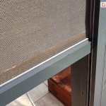 Zdjęcie prezentuje szczegółowe zbliżenie na pionowy refleksol Zip zamontowany w konstrukcji pergoli. Widoczna jest techniczna tkanina o gęstym splocie w odcieniach brązu i beżu, która zapewnia ochronę przed słońcem i wiatrem. Dolna krawędź materiału wykończona jest estetyczną, aluminiową listwą obciążającą w kolorze srebrzystografitowym, która stabilizuje roletę podczas jej rozwijania. Tkanina porusza się precyzyjnie wewnątrz ciemnej, antracytowej prowadnicy bocznej zintegrowanej ze słupem pergoli. W tle, pod roletą, widoczne są fragmenty tarasu wyłożonego deskami oraz białe kamienie ozdobne, co podkreśla nowoczesny i zadbany charakter otoczenia.