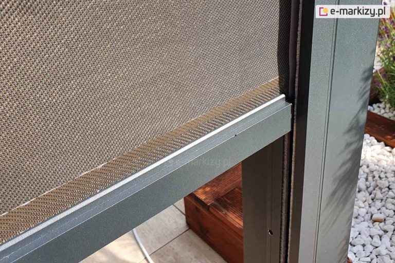 Zdjęcie prezentuje szczegółowe zbliżenie na pionowy refleksol Zip zamontowany w konstrukcji pergoli. Widoczna jest techniczna tkanina o gęstym splocie w odcieniach brązu i beżu, która zapewnia ochronę przed słońcem i wiatrem. Dolna krawędź materiału wykończona jest estetyczną, aluminiową listwą obciążającą w kolorze srebrzystografitowym, która stabilizuje roletę podczas jej rozwijania. Tkanina porusza się precyzyjnie wewnątrz ciemnej, antracytowej prowadnicy bocznej zintegrowanej ze słupem pergoli. W tle, pod roletą, widoczne są fragmenty tarasu wyłożonego deskami oraz białe kamienie ozdobne, co podkreśla nowoczesny i zadbany charakter otoczenia.