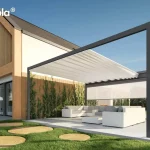Wizualizacja przedstawia nowoczesną pergolę tarasową Tarasola Flat o prostej, geometrycznej bryle, zamontowaną przy budynku o architekturze typu nowoczesna stodoła. Konstrukcja pergoli w kolorze ciemnego antracytu posiada jasne, kremowe zadaszenie tkaninowe, które jest częściowo rozsunięte, tworząc estetyczne fale podwieszone na poprzecznych profilach. Na jednym z boków konstrukcji zainstalowano białą roletę zewnętrzną typu screen, która jest w połowie rozwinięta, osłaniając strefę wypoczynku. Pod pergolą znajduje się duży, narożny zestaw mebli ogrodowych w kolorze białym z jasnoszarymi poduszkami oraz pasujący stolik kawowy. Całość usytuowana jest na jasnym, wielkoformatowym tarasie, który sąsiaduje z zadbanym trawnikiem z dekoracyjnymi, okrągłymi płytami chodnikowymi imitującymi przekrój pnia drzewa. Elewacja domu łączy biały tynk z pionowymi lamelami drewnianymi, a przy ścianie widoczne są pnące rośliny, co podkreśla luksusowy i naturalny charakter aranżacji.