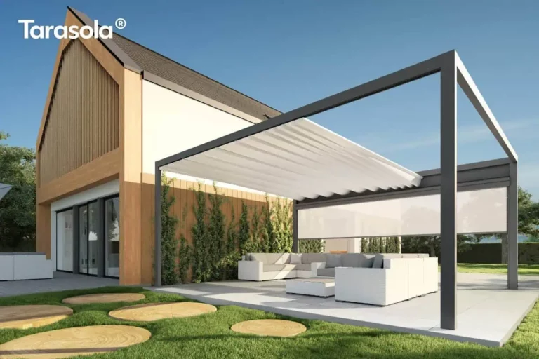 Wizualizacja przedstawia nowoczesną pergolę tarasową Tarasola Flat o prostej, geometrycznej bryle, zamontowaną przy budynku o architekturze typu nowoczesna stodoła. Konstrukcja pergoli w kolorze ciemnego antracytu posiada jasne, kremowe zadaszenie tkaninowe, które jest częściowo rozsunięte, tworząc estetyczne fale podwieszone na poprzecznych profilach. Na jednym z boków konstrukcji zainstalowano białą roletę zewnętrzną typu screen, która jest w połowie rozwinięta, osłaniając strefę wypoczynku. Pod pergolą znajduje się duży, narożny zestaw mebli ogrodowych w kolorze białym z jasnoszarymi poduszkami oraz pasujący stolik kawowy. Całość usytuowana jest na jasnym, wielkoformatowym tarasie, który sąsiaduje z zadbanym trawnikiem z dekoracyjnymi, okrągłymi płytami chodnikowymi imitującymi przekrój pnia drzewa. Elewacja domu łączy biały tynk z pionowymi lamelami drewnianymi, a przy ścianie widoczne są pnące rośliny, co podkreśla luksusowy i naturalny charakter aranżacji.