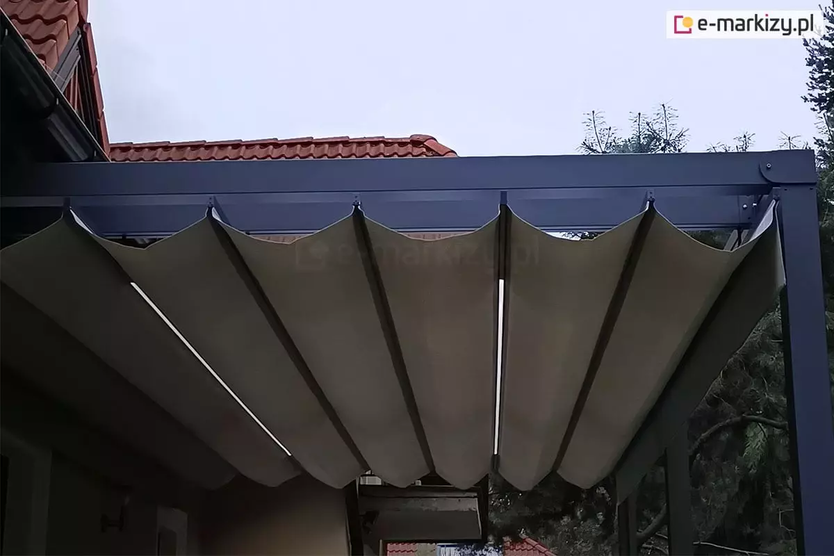 Zbliżenie od dołu na dach tkaninowy rzymski Tako w trakcie rozkładania. Zdjęcie przedstawia charakterystyczne, głębokie fałdy materiału technicznego w kolorze jasnobeżowym, które są podwieszone na poprzecznych profilach usztywniających. W dwóch belkach przeciwwietrznych zastosowano taśmy ledowe. Profile te poruszają się wewnątrz aluminiowej prowadnicy bocznej w kolorze antracytowym. Konstrukcja jest zamontowana do ściany budynku (widoczny fragment jasnej elewacji po lewej) i wsparta na słupach pionowych. W górnej części kadru widoczny jest fragment dachu budynku z ceramiczną dachówką w kolorze ceglastym. Tło stanowią korony ciemnych drzew iglastych oraz jasne niebo. Perspektywa zdjęcia podkreśla symetrię systemu fałdowania tkaniny oraz solidność antracytowej ramy konstrukcyjnej.