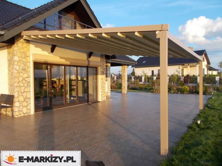 Nowoczesna przyścienna pergola tarasowa o aluminiowej konstrukcji w kolorze piaskowego beżu, zamontowana do elewacji piętrowego domu wykończonego jasnym tynkiem i ozdobnym kamieniem elewacyjnym. Zadaszenie posiada ruchomy system tkaninowy w kolorze kremowym, który jest częściowo rozsunięty, odsłaniając niebo. Konstrukcja wspiera się na dwóch solidnych, pionowych słupach o kwadratowym przekroju, posadowionych na rozległym tarasie wyłożonym ciemnobrązowymi płytkami gresowymi o strukturze drewna. Pod pergolą widoczne są duże, przeszklone drzwi przesuwne prowadzące do wnętrza salonu, nad którymi zamontowano zewnętrzną roletę. W tle rozpościera się zadbany ogród z niską roślinnością oraz widok na sąsiednie budynki jednorodzinne o podobnej architekturze. Zdjęcie wykonane w ciepłym, popołudniowym świetle słonecznym, prezentuje szeroki kąt zagospodarowania przestrzeni rekreacyjnej przy nowoczesnym domu.