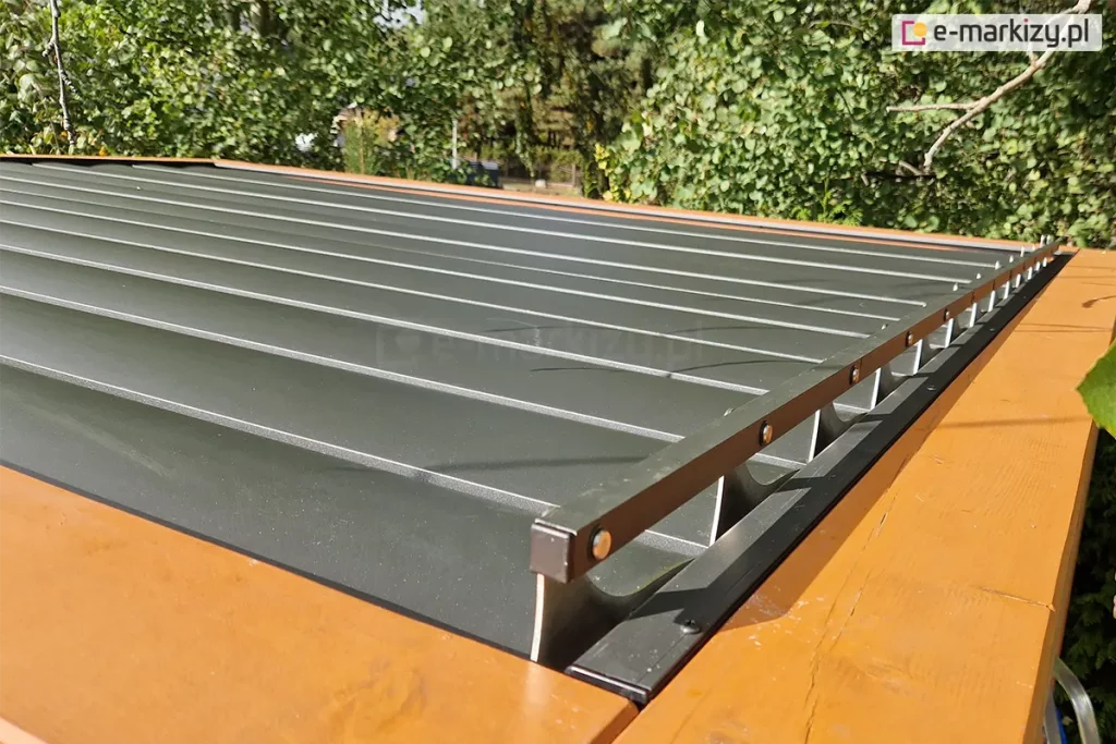 Drewniana konstrukcja z zamocowanym na niej dachem lamelowym SB400 Selt z otwartymi lamelami o antracytowym kolorze. Na pierwszym planie widoczny mechanizm sterowania ruchem lameli. W tle widoczne drzewa liściaste.