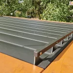 Drewniana konstrukcja z zamocowanym na niej dachem lamelowym SB400 Selt z otwartymi lamelami o antracytowym kolorze. Na pierwszym planie widoczny mechanizm sterowania ruchem lameli. W tle widoczne drzewa liściaste.