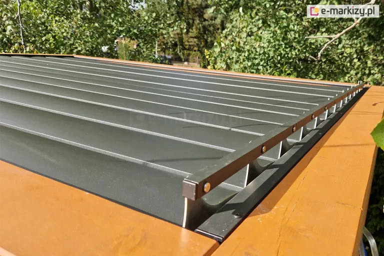 Drewniana konstrukcja z zamocowanym na niej dachem lamelowym SB400 Selt z otwartymi lamelami o antracytowym kolorze. Na pierwszym planie widoczny mechanizm sterowania ruchem lameli. W tle widoczne drzewa liściaste.