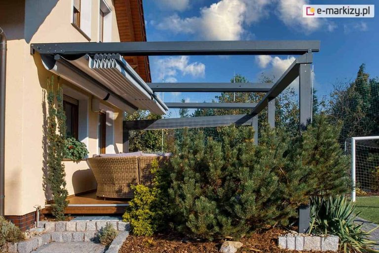 widok z boku na nowoczesną pergolę aluminiową Solid w wersji przyściennej, zamontowaną nad drewnianym tarasem. Konstrukcja w kolorze ciemnego antracytu posiada dach tkaninowy, który na tym ujęciu jest całkowicie zwinięty w kierunku elewacji budynku, co odsłania widok na niebo i wpuszcza światło do wnętrza domu. Na tarasie znajduje się duży, półokrągły narożnik wykonany z technorattanu z jasnymi poduszkami. Otoczenie pergoli charakteryzuje się bogatą, zadbaną roślinnością – na pierwszym planie widać rozłożystą sosnę górską oraz pnące iglaki przy ścianie budynku. Dom ma jasną, żółtawą elewację, a w oddali, po prawej stronie, widoczna jest zielona przestrzeń ogrodu z bramką piłkarską.