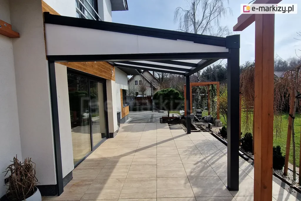 Zadaszenie tarasu Modes z wypełnieniem z poliwęglanu i antracytową konstrukcją, uzupełnione o boczny trójkąt z poliwęglanu. Całość ustawiona na tarasie z płyt gresowych przed domkiem jednorodzinnym o szerokim oknie tarasowym. W tle widoczne rośliny ogrodowe, drzewka i trawnik, a także sąsiedni budynek.