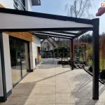 Zadaszenie tarasu Modes z wypełnieniem z poliwęglanu i antracytową konstrukcją, uzupełnione o boczny trójkąt z poliwęglanu. Całość ustawiona na tarasie z płyt gresowych przed domkiem jednorodzinnym o szerokim oknie tarasowym. W tle widoczne rośliny ogrodowe, drzewka i trawnik, a także sąsiedni budynek.